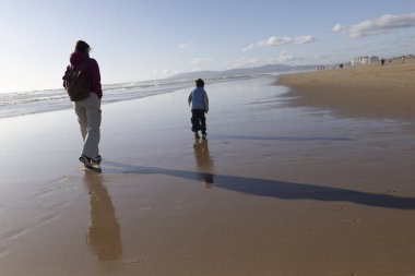 Anne ve çocuk gün batımında ıslak kumlu bir sahilde yürüyorlar, uzun gölgeler bırakarak.