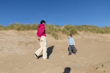 Anne ve oğlu güneşli bir günde çimlerle kaplı kum tepelerinin yakınındaki kumsalda birlikte oynuyorlar.