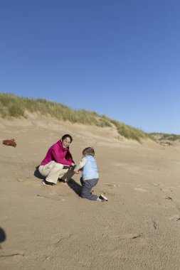 Anne ve oğlu, güneşli bir sahilde kumdan kaleler inşa ederek güzel vakit geçiriyorlar.