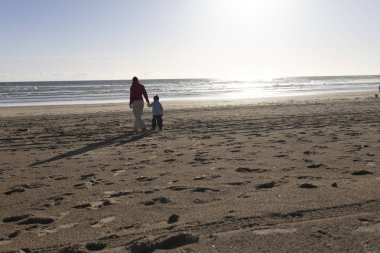 Anne ve oğlu gün batımında kumlu bir plajda rahatça geziniyorlar.