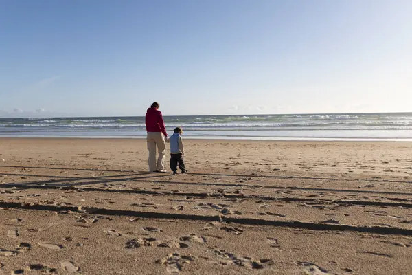 Anne ve oğul el ele tutuşuyor, okyanusun kıyısındaki kumsalda rahatlatıcı bir yürüyüşün tadını çıkarıyorlar.