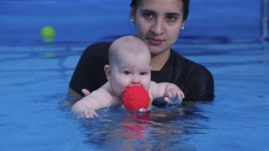 Yüzme havuzunda kırmızı topu tutan bebeğe yardımcı olan eğitmen.