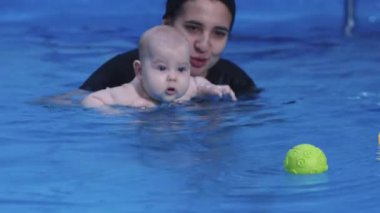 Genç anne bebeğine kapalı havuzda yardım ediyor ve bebeği suya sokuyor.