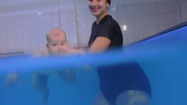 Kapalı havuzda yüzme dersleri sırasında bir bebeğe yardım eden eğitmen.