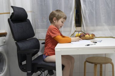 Ödev yapan küçük bir çocuk, büyük bir ofis sandalyesinde oturuyor ve mutfak masasına yerleştirilmiş bir defter çiziyor.