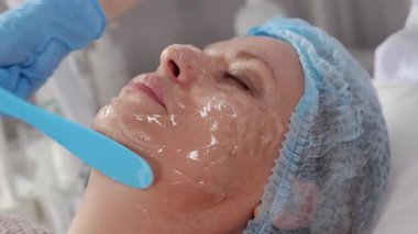 Kozmetik uzmanı kadın müşterinin yüzüne nemlendirici jel maske yayıyor.