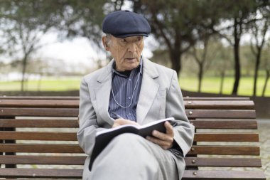 Yaşlı bir adam bankta kulaklıkla müzik dinlerken deftere notlar alıyor.