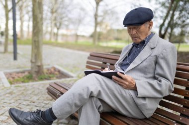 Takım elbiseli ve şapkalı yaşlı bir adam, parkta bankta oturuyor, kulaklıkla müzik dinliyor ve defterde yazıyor.
