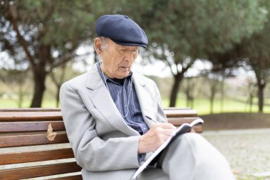 Not defterine not alan, kulaklık takan, müzik dinleyen, bankta oturan kıdemli bir adam.