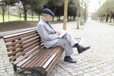Yaşlı profesör, parktaki bankta kulaklıkla müzik dinlerken deftere notlar alıyor.