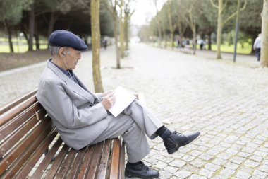 Takım elbiseli ve bereli yaşlı bir adam bankta oturuyor, notlar alıyor ve kulaklıkla müzik dinliyor.