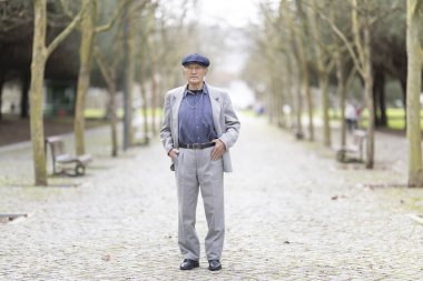 Kendinden emin yaşlı bir adam ağaçlarla kaplı bir kaldırım yolunda duruyor, elleri ceplerinde poz veriyor.
