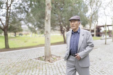 Takım elbiseli ve düz şapkalı bir adam parkta yürüyor.