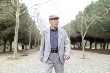 Takım elbiseli ve şapkalı yaşlı bir adam parkta yürürken müzik dinliyor.