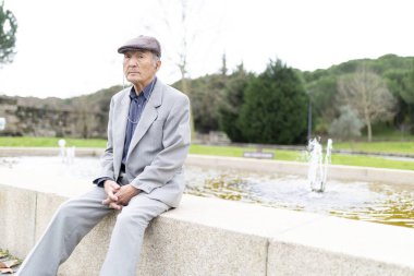 Takım elbiseli ve yassı şapkalı bir adam huzur dolu bir park ortamında çeşmenin yanında dinleniyor.