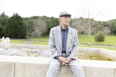 Takım elbiseli ve yassı şapkalı bir adam, sakin bir park ortamındaki çeşmenin kenarında derin derin otururken gözlerini kaçırıyor.