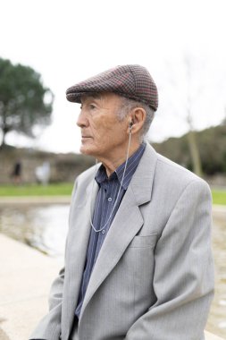 Huzurlu bir park ortamında otururken kulaklıkla müzik dinlemeyi seven kıdemli bir adam.