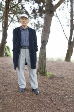 Parkta çam ağaçlarıyla poz veren şık kıdemli adam aktif yaşlanma ve doğanın tadını çıkartıyor.