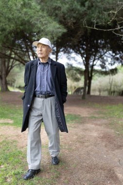 Kıdemli Asyalı bir adam parkta rahatça yürüyüp, şapka takıp müzik dinliyor.