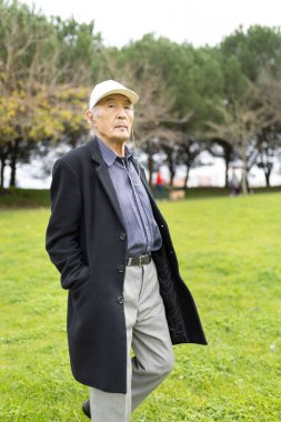 Kıdemli Asyalı adam yeşil bir parkta yürürken kulaklıkla müzik dinliyor.