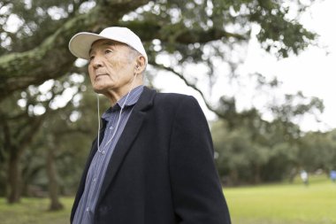 Kıdemli Asyalı adam yeşil bir parkta dururken kulaklık takıyor ve müzikten zevk alıyor.