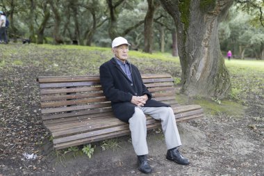 Siyah ceketli ve beyaz şapkalı yaşlı bir adam ahşap bir bankta oturuyor, ağaçların gölgesinde huzurlu atmosferin tadını çıkarıyor.