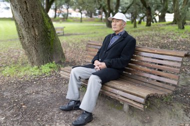 Yaşlı Asyalı kulaklık takan bir adam parkta bankta oturmuş, müzik ve temiz havanın tadını çıkarıyor.