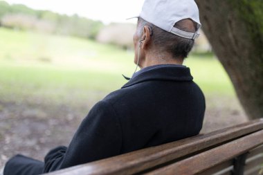 İşitme cihazı olan yaşlı bir adam parktaki bankta oturmuş doğanın tadını çıkarıyor.