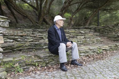 Huzurlu bir park ortamında taş basamaklarda otururken kulaklıkla müzik dinleyen yaşlı bir adam.