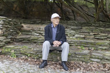 Kıdemli adam, doğayla çevrili bir parkta taş basamaklarda otururken huzur dolu bir anın tadını çıkarıyor.