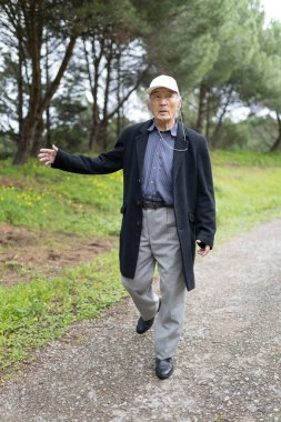 Yaşlı profesör bir ceket ve şapka giyip parkta yürüyor ve eliyle el kol hareketi yapıyor.