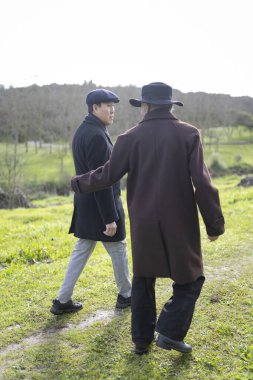 Zarif paltolar giyen iki işadamı yeşil bir alanda yürüyor ve konuşuyorlar, doğanın ortasında bir anlık bağlantının tadını çıkarıyorlar.