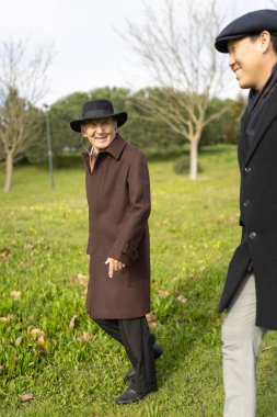 Son sınıf öğrencisi şapkalı ve bakıcı parkta birlikte yürüyor, sohbetin ve temiz havanın tadını çıkarıyor.