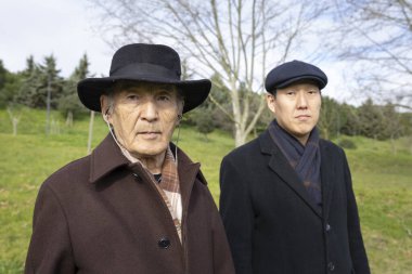 Paltolu ve şapkalı Asyalı baba ve oğul aile bağlarını ve nesillerarası bağlantıyı temsil eden bir parkta yan yana duruyorlar.