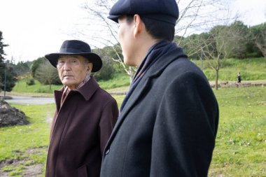 Asyalı baba ve oğul zarif paltolar giyip parkta yürüyüp sohbet ediyorlar.