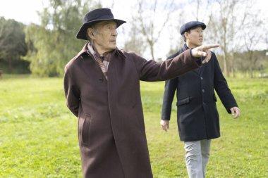 Yaşlı bir adam, genç bir adamla dışarıda yürürken uzaktaki bir şeyi işaret ediyor. Parkta rahatça geziniyor.