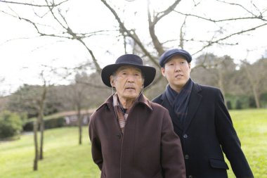 Asyalı yetişkin oğul ve yaşlı baba birlikte vakit geçiriyorlar. Açık havada parkta kaynaşıyorlar.