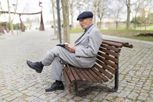 Yaşlı bir adam bankta kulaklıkla müzik dinlerken defterine notlar alıyor.