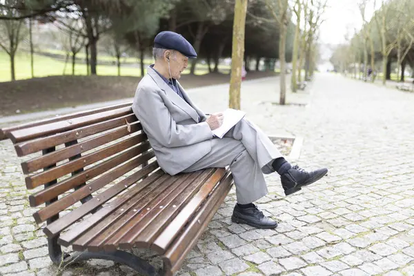 Yaşlı profesör, parktaki bankta kulaklıkla müzik dinlerken deftere notlar alıyor.