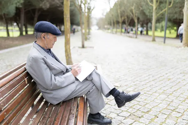 Takım elbiseli ve bereli yaşlı bir adam bankta oturuyor, notlar alıyor ve kulaklıkla müzik dinliyor.