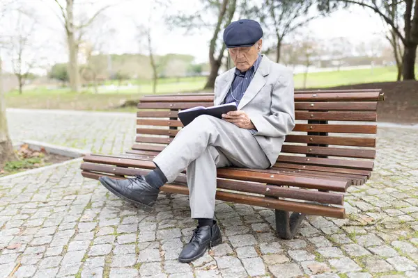 Huzurlu bir anın tadını çıkaran yaşlı bir beyefendi kitap okuyor ve parkta otururken müzik dinliyor.