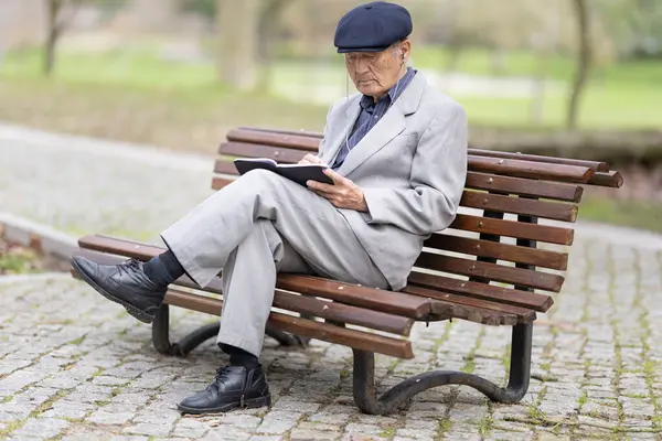 Park bankında otururken deftere yazı yazan kıdemli yazar, huzurlu bir ortamın tadını çıkarıyor.