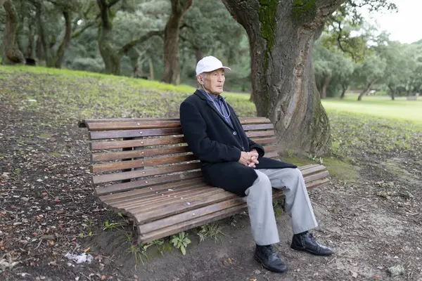 Siyah ceketli ve beyaz şapkalı yaşlı bir adam sonbahar sezonunda parktaki ahşap bir bankta oturuyor.