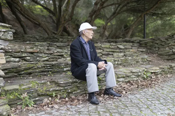 Huzurlu bir park ortamında taş basamaklarda otururken kulaklıkla müzik dinleyen yaşlı bir adam.