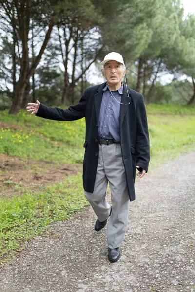 Yaşlı profesör bir ceket ve şapka giyip parkta yürüyor ve eliyle el kol hareketi yapıyor.