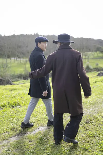 Zarif paltolar giyen iki işadamı yeşil bir alanda yürüyor ve konuşuyorlar, doğanın ortasında bir anlık bağlantının tadını çıkarıyorlar.