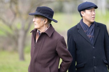 Şapka ve ceket giyen iki kıdemli Asyalı parkta duruyor, muhtemelen sohbet ediyorlar ya da manzaranın tadını çıkarıyorlar.