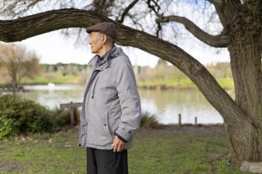 Huzurlu bir park ortamında gölün kenarında duran gri ceketli ve düz şapkalı yaşlı adam.