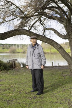 Huzurlu bir parkta gölün kenarında duran yaşlı adam, doğanın huzurunun tadını çıkarıyor.