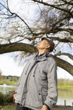 Yaşlı bir adam gölün yakınındaki bir parkta ağaçlara bakarken kulaklıkla müzik dinliyor.